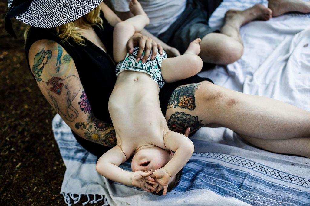 A tattooed person wearing a hat sits on a picnic blanket, holding a baby wearing a diaper with a green pattern. The baby is lying on their back with arms covering their face. Another person’s leg is partially visible. Trees surround them.