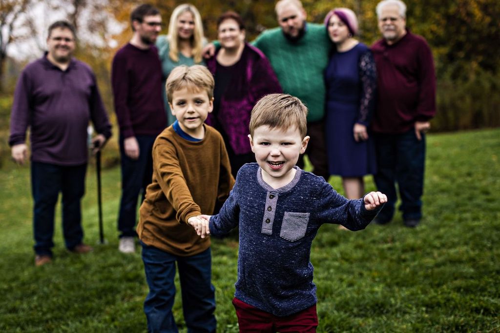 Two young boys in the foreground are smiling and running towards the camera. Behind them, a group of five adults and one elderly person stand together on a grassy area with trees in the background. The scene is cheerful and outdoor.
