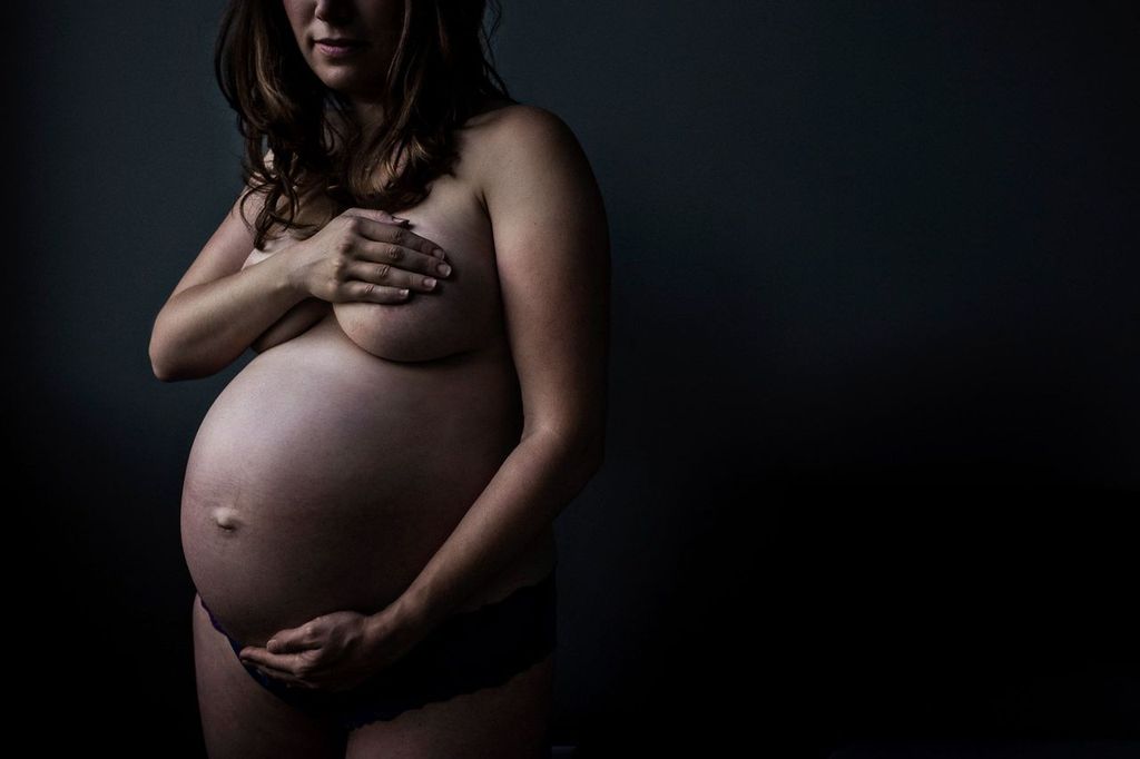 A pregnant woman stands against a dark background, holding her belly with one hand and covering her chest with the other. Her expression is calm and serene. The lighting emphasizes her baby bump and creates a soft, intimate atmosphere.