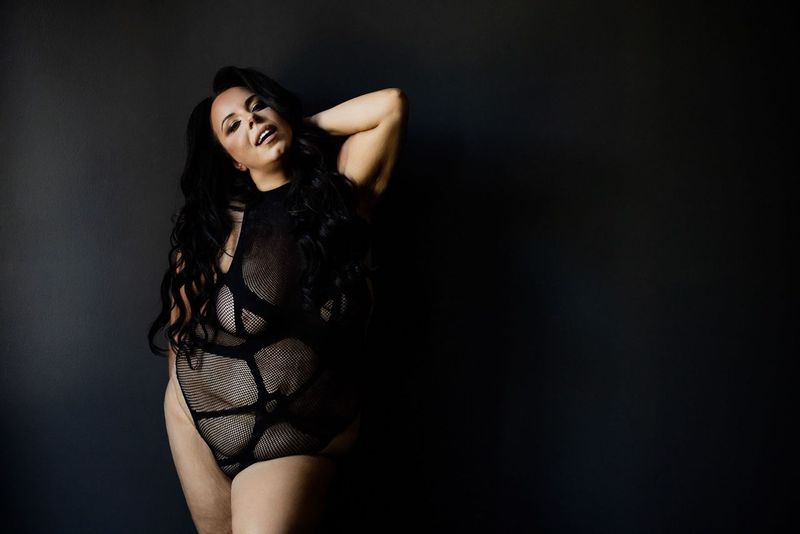 Woman with long dark hair wearing a black, patterned bodysuit poses confidently against a dark background, smiling with one hand behind their head.