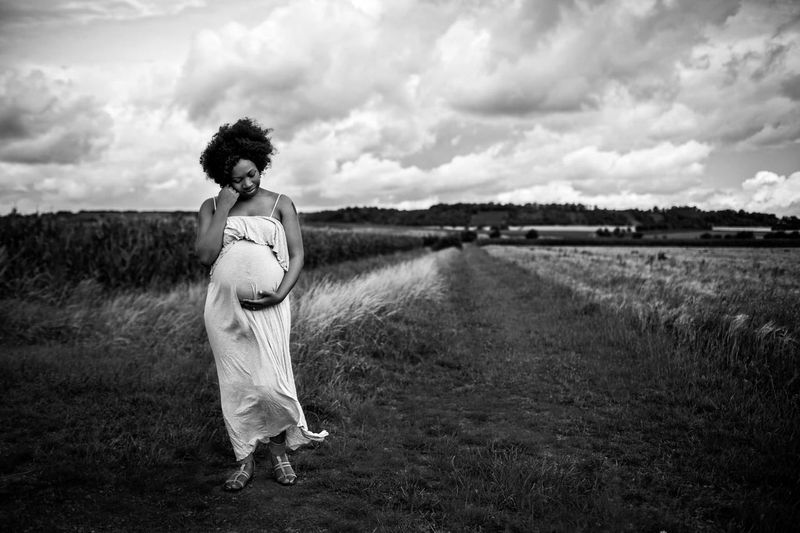 A pregnant woman stands on a dirt path in a field, cradling her belly and looking down thoughtfully. She wears a long, flowing dress. The background features tall grasses and a cloudy sky, creating a serene and contemplative atmosphere.