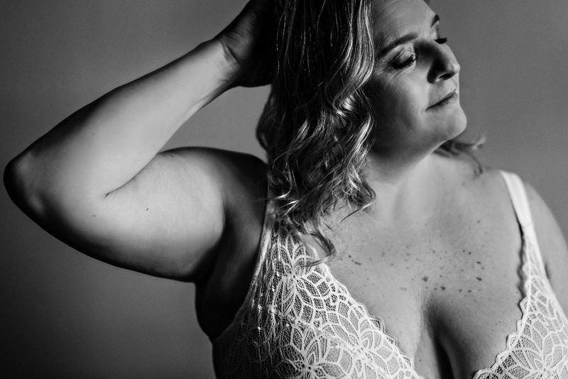 Black and white portrait of a woman with wavy hair, wearing a lace top. Her eyes are gently closed as she tilts her head back slightly, one hand raised to her hair, creating a serene and contemplative mood.