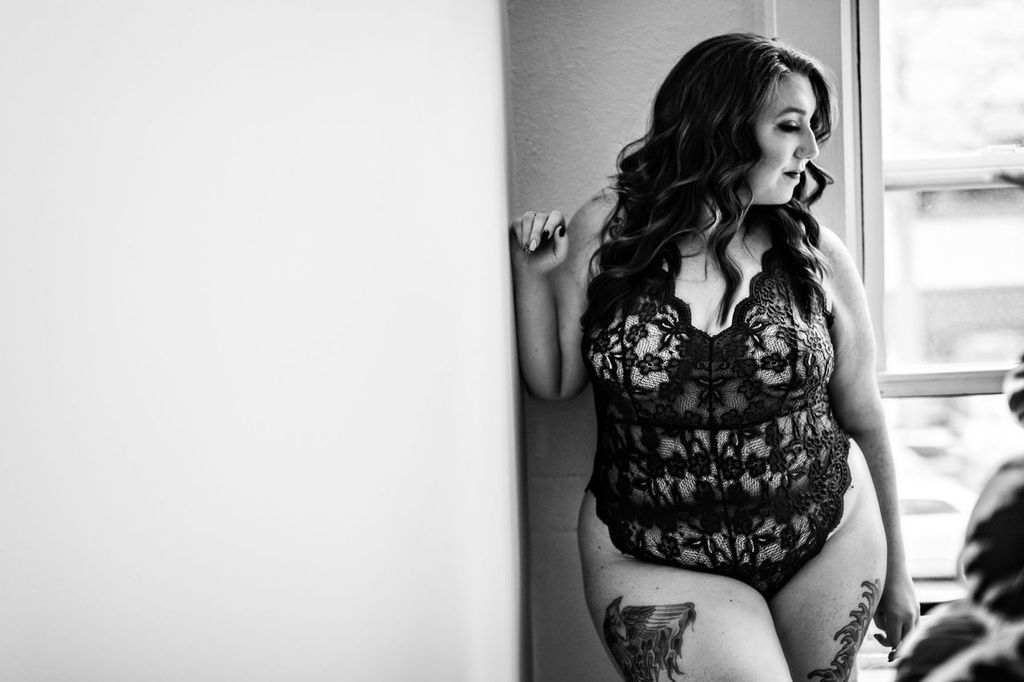 A woman with wavy hair, wearing a black lace bodysuit, leans against a wall by a window. She has tattoos on her thigh and arm. The black and white photo captures her looking thoughtfully outside.