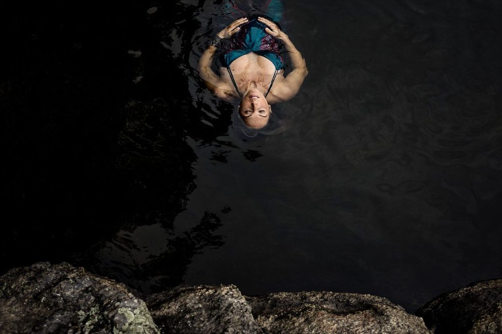 A person with long hair floats peacefully on their back in dark, calm water. They are wearing a blue swimsuit, and their eyes are closed. Rock formations are visible at the bottom of the image.