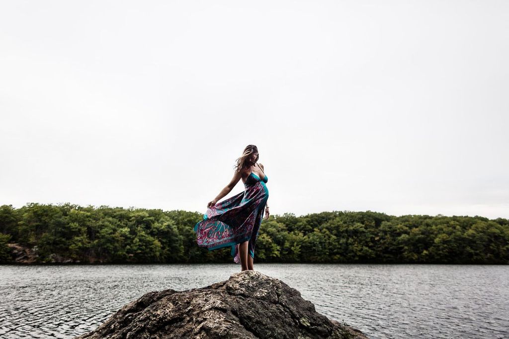 A woman stands on a rock by the water, holding a flowing, colorful dress. She faces away, looking at a lush green forest across the lake under a cloudy sky.