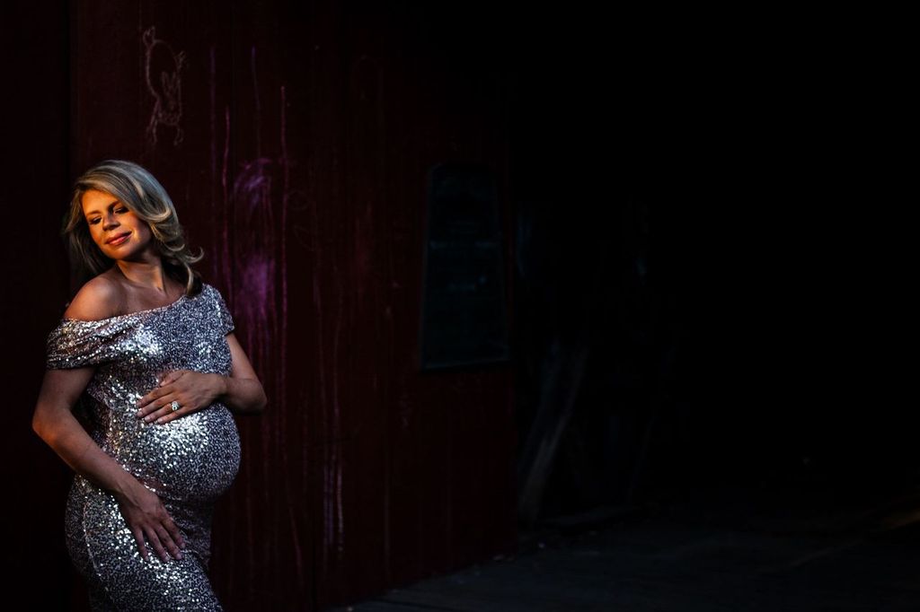 A pregnant woman in a sparkling, off-the-shoulder dress poses contentedly, cradling her belly, against a dark backdrop with faint red streaks. She is illuminated by soft light, highlighting the dresss sequins.