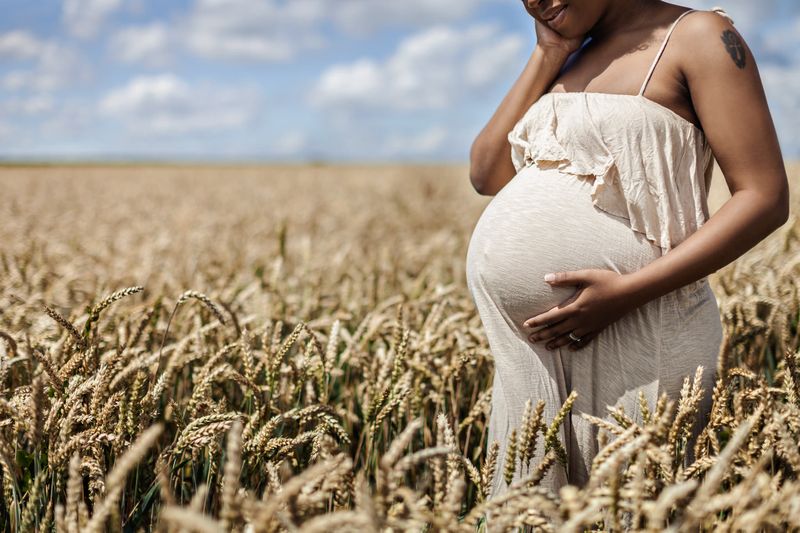 A pregnant woman in a light dress stands in a field of wheat, gently cradling her belly with one hand and touching her face with the other. The sky is blue with scattered clouds, creating a serene and peaceful scene.