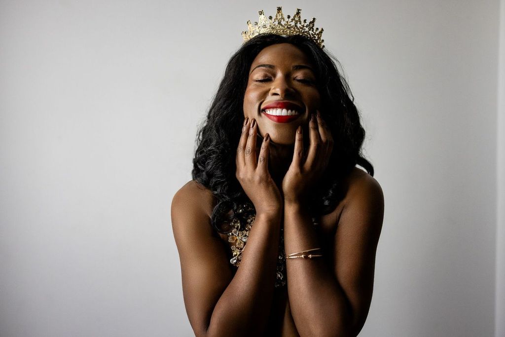 A person with long black hair smiles joyfully with eyes closed, wearing a gold crown and a bejeweled necklace. Their hands are gently touching their face, exuding a sense of happiness and confidence.