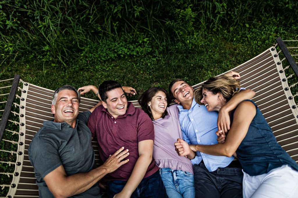 Family of five people are laughing and lying side by side on a hammock in a garden. The family includes two women and three men, dressed casually, and enjoying a lighthearted moment together. Lush greenery surrounds them.