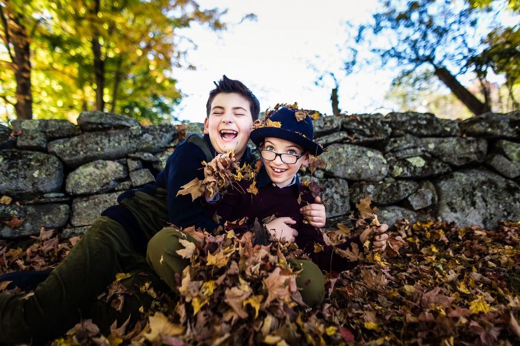 Two children joyfully play in a pile of autumn leaves near a stone wall. One child, wearing a black jacket, laughs while reclining in the leaves. The other, wearing glasses and a navy hat, smiles and throws leaves. Trees with yellow leaves are in the background.