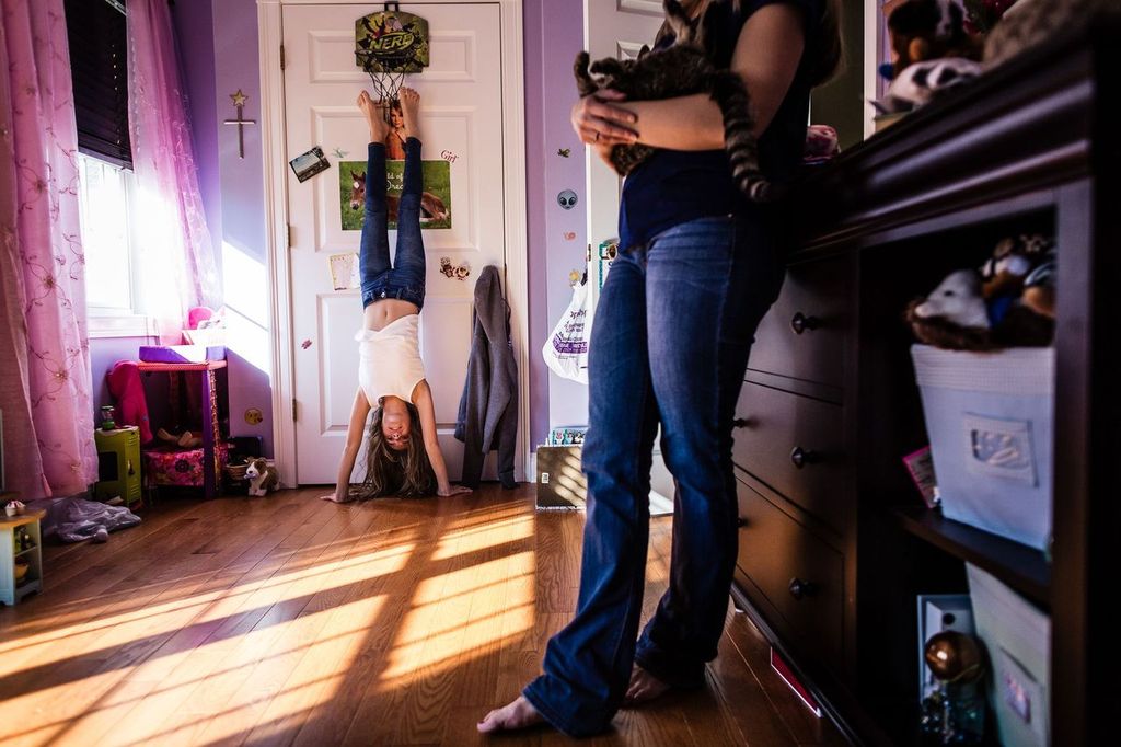 A girl performs a handstand against a closed door in a colorful room while another person, standing nearby, holds a cat. The room is decorated with various items, toys, and pink curtains, and sunlight casts shadows on the wooden floor.
