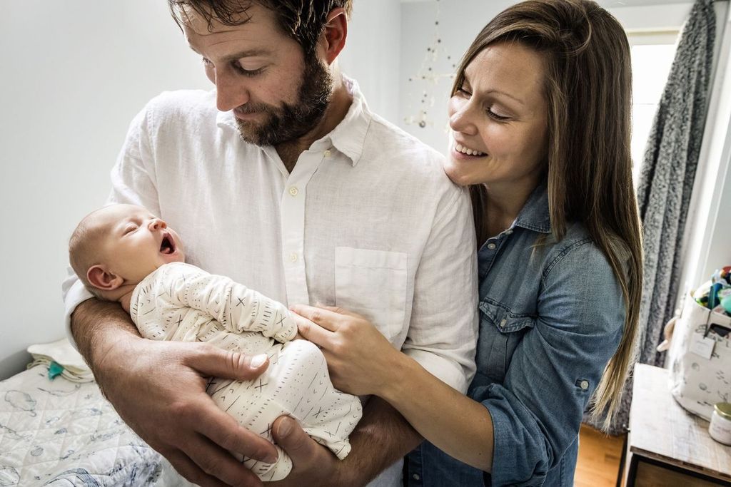 A smiling couple is in a softly lit room, gazing lovingly at a yawning baby wrapped in a patterned onesie. The man cradles the baby while the woman gently touches the babys arm. A crib is visible in the background.