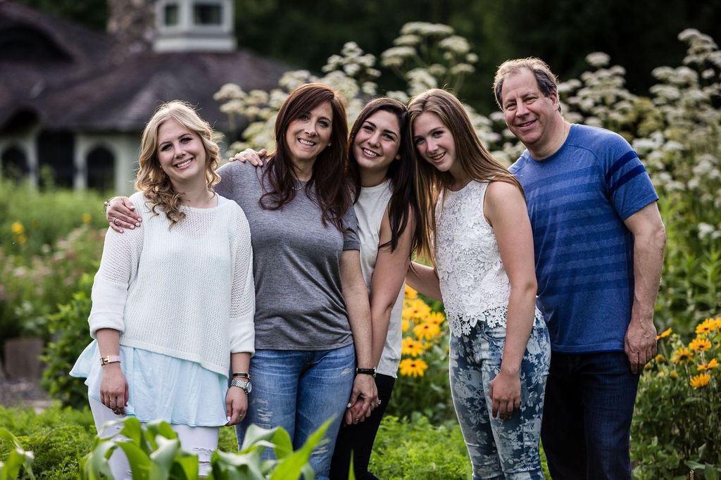A family of five people stand closely together outdoors, smiling in front of a background filled with green foliage and white flowers. Four women and one man are enjoying a sunny day in casual clothing.