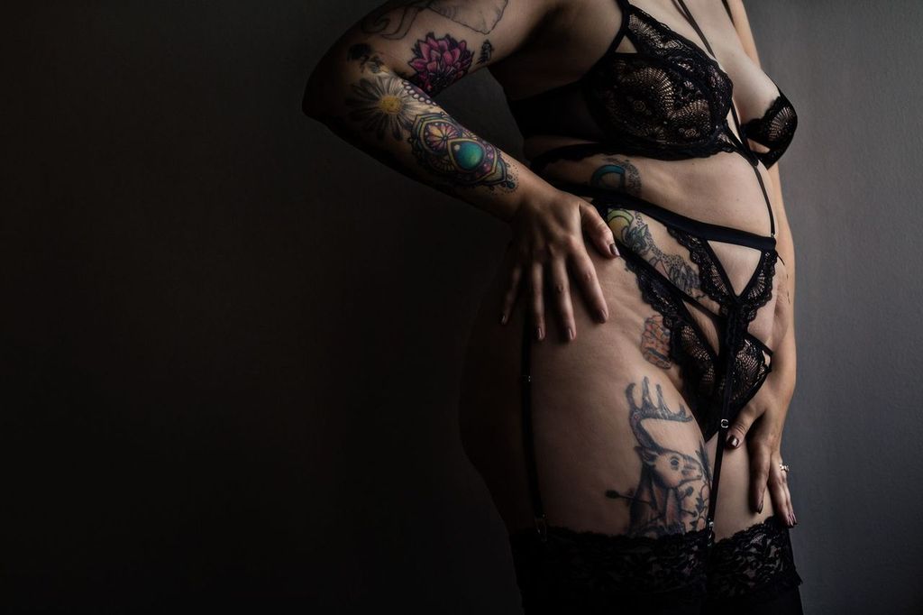A person wearing black lingerie poses against a dark background, showcasing tattoos on their arms, torso, and thigh. Their face is not visible, and light highlights their tattoos and attire.