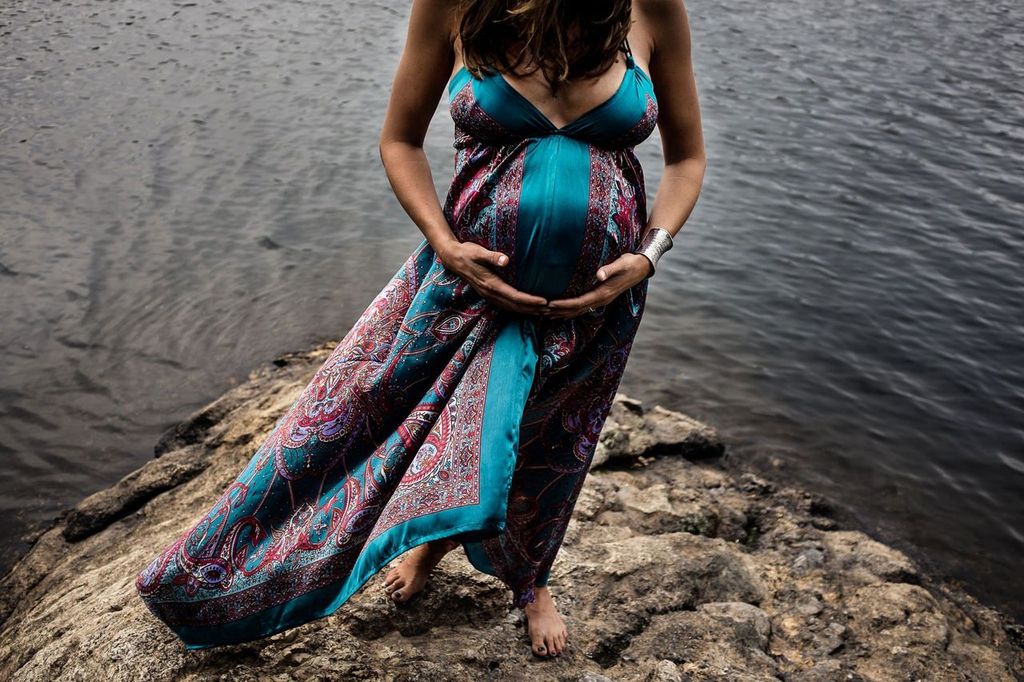 Pregnant woman in a colorful, flowing dress stands barefoot on a rock by the water, gently cradling her belly with both hands. Her hair falls loosely, and she wears a silver bracelet, conveying a serene, natural setting.