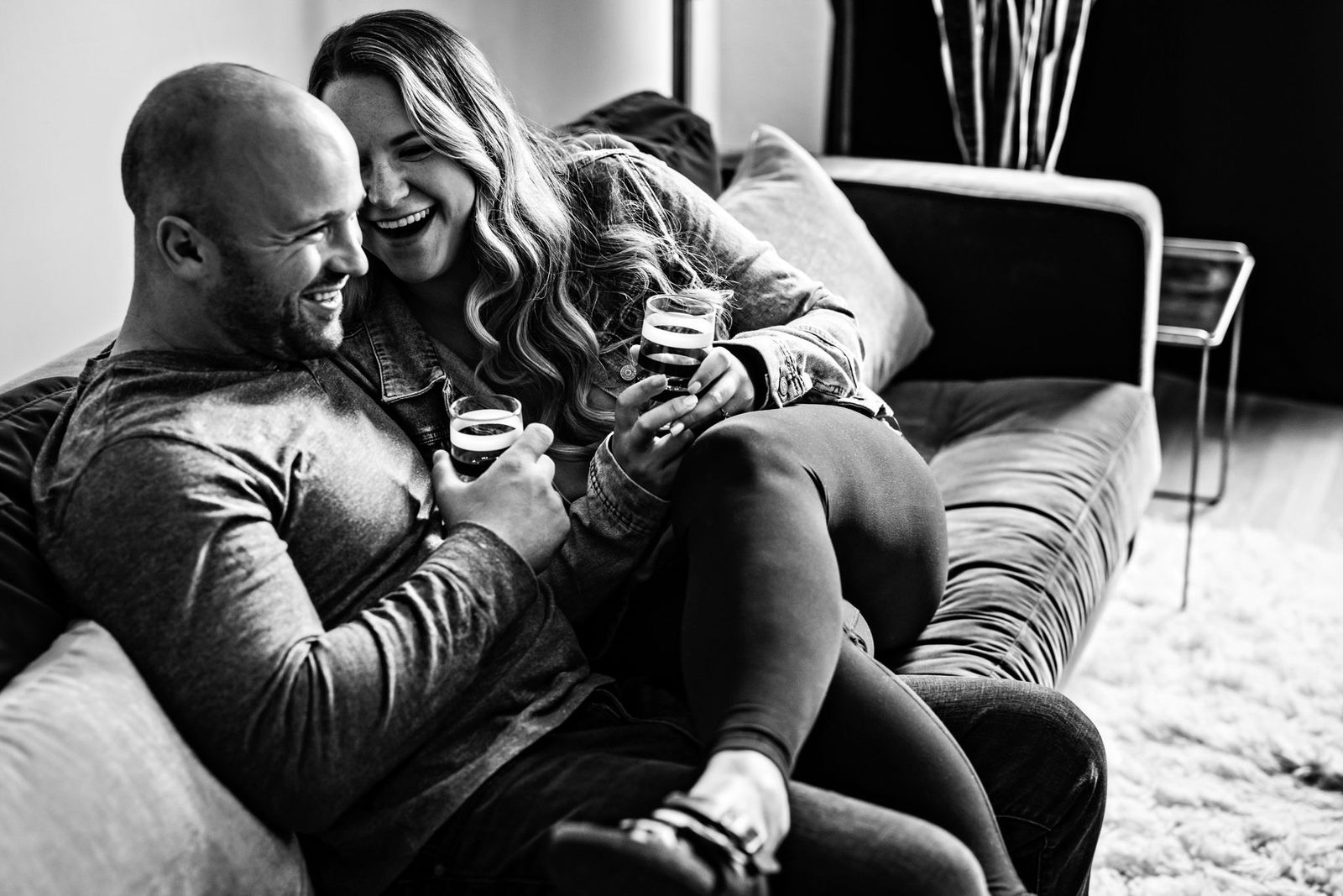 A black and white image of a smiling couple sitting close together on a sofa, holding cups. The woman is leaning into the man, both appear relaxed and happy. Soft lighting creates a cozy atmosphere.