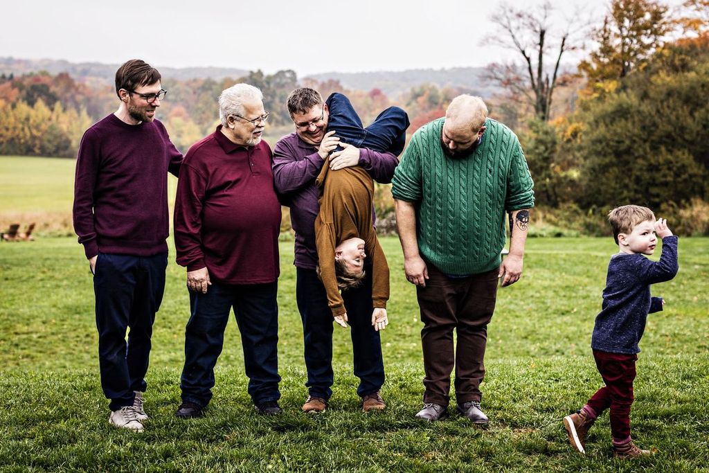 A group of five adults and one child are outdoors on grass. One adult is playfully holding the child upside down. Another child is seen running with his hand over his eyes, while the adults are dressed in various sweaters and appear amused. Trees in the background.