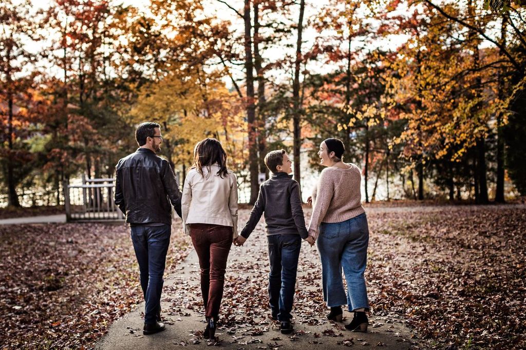 Four people walk hand in hand down a leaf-covered path in a park during autumn. They are wearing casual clothing, and the trees around them have vibrant fall foliage.