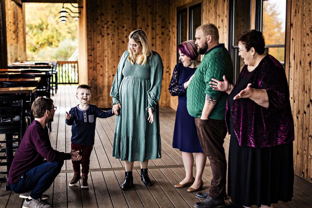 A family gathers on a wooden porch, featuring a smiling child with a man crouched beside him, and four adults standing nearby. The group appears relaxed and happy, engaged in conversation with each other.