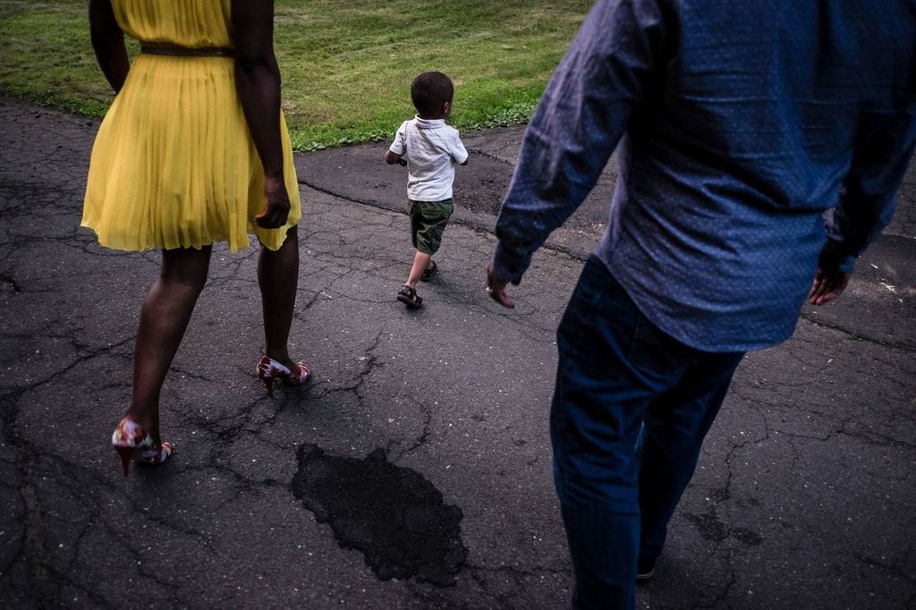 A child wearing a white shirt and green shorts walks on a cracked asphalt path, flanked by two adults. The adult on the left wears a yellow dress, and the one on the right wears a blue shirt. Grass is visible in the background.