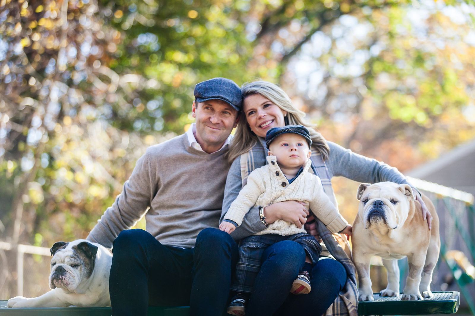 A family with a man, woman, and a child wearing hats sit outdoors on a bench, flanked by two bulldogs. They are in a park, surrounded by trees with autumn foliage. Everyone is smiling and dressed in cozy, neutral-colored clothing.