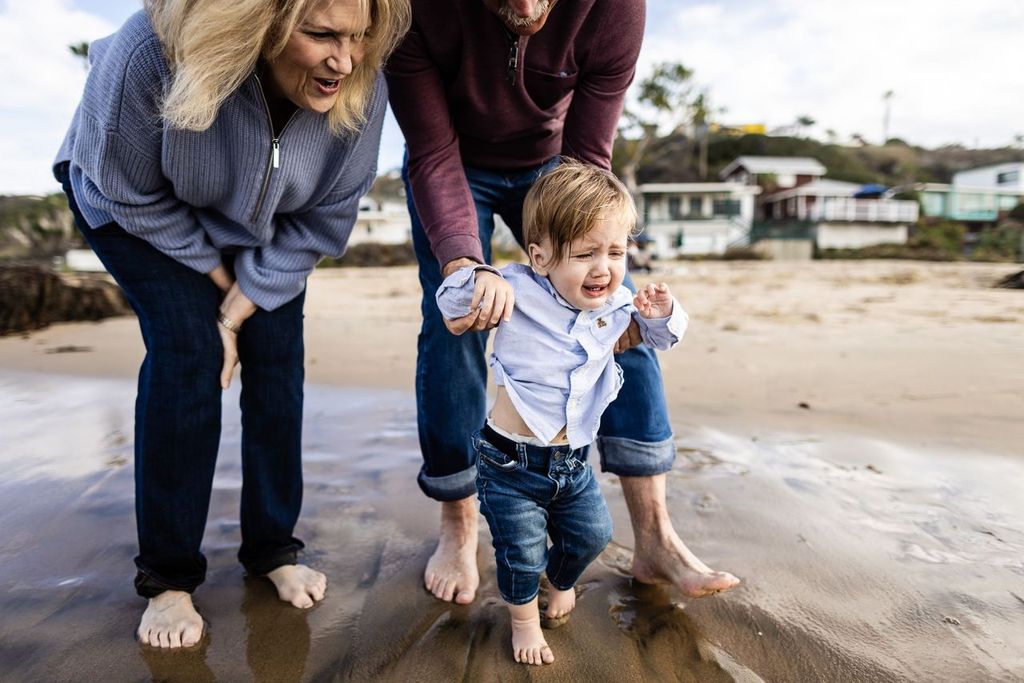 A small child walks barefoot on the beach with the assistance of two adults. One adult holds the childs arm to support. The child is wearing jeans and a partially buttoned shirt, while the adults wear sweaters and jeans.