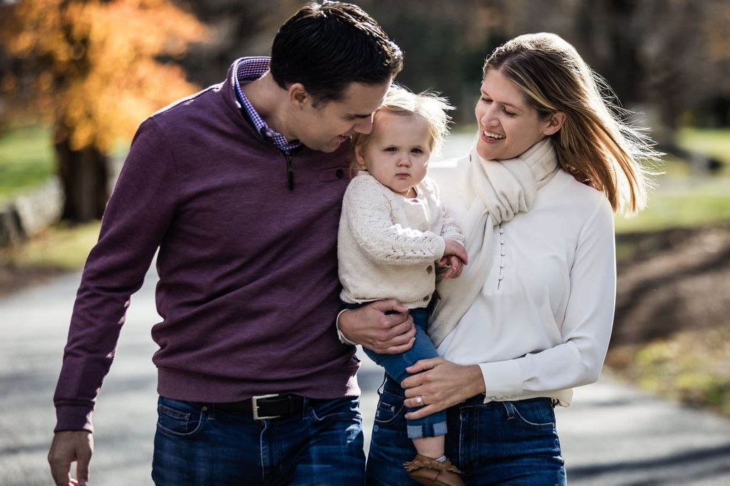A couple walks along a path, holding a young child between them. The man wears a maroon sweater, and the woman is in a white top and scarf. The child looks at the camera with a neutral expression. Autumn trees are in the background.