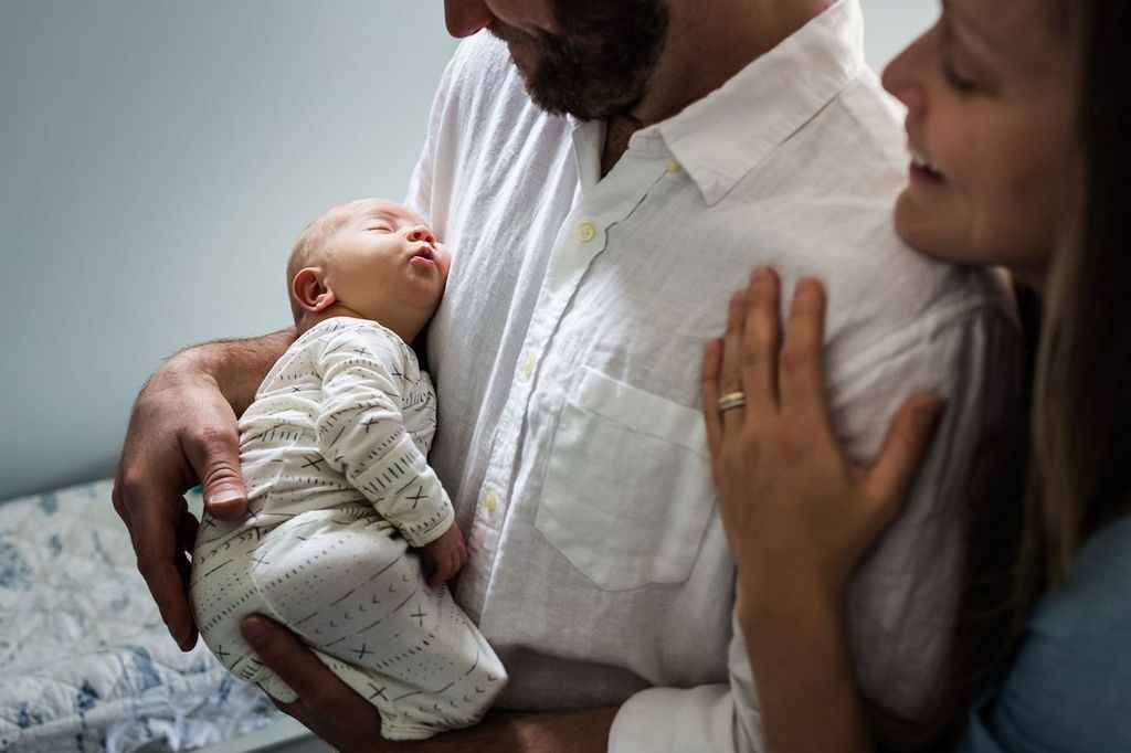 A baby sleeps peacefully in the arms of father wearing a white shirt, while the mother stands close, smiling with a hand on the shoulder of the father. The scene conveys warmth and tenderness.