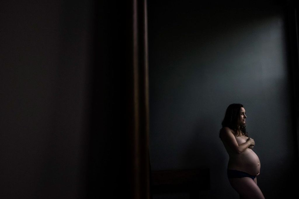 A pregnant woman stands in profile, holding her belly, in a dimly lit room. The focus is on her, highlighting the contours of her silhouette against the dark, muted background.