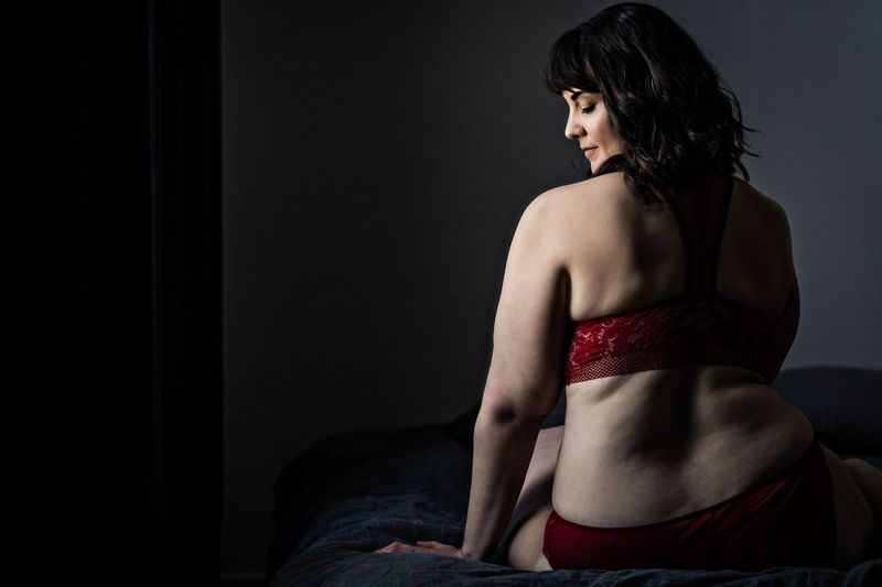 A woman with dark hair sits on a bed, wearing red lingerie. She is turned slightly away from the camera, with soft lighting illuminating her back and shoulders. The room is dimly lit, creating a serene and intimate atmosphere.