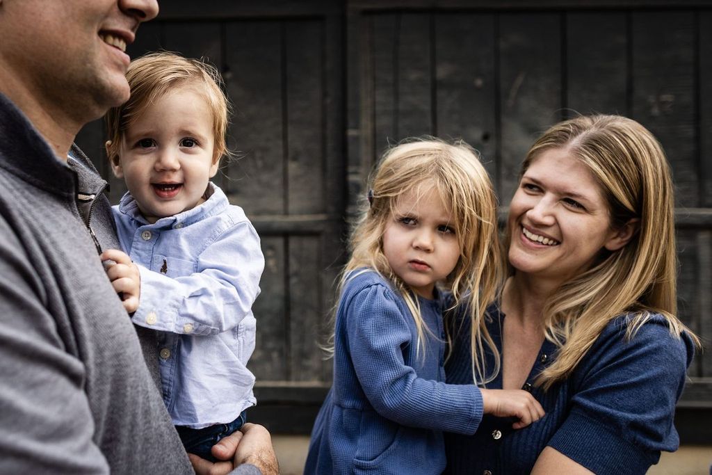 A family is gathered outdoors against a wooden background. A man holds a smiling toddler boy in a blue shirt, while a woman with long blonde hair is holding a young girl in a blue dress. The woman is smiling at the girl.