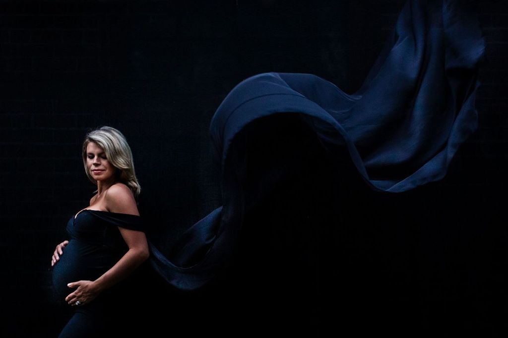 Pregnant woman in a flowing black dress poses against a dark background, gently holding her belly. The dress fabric billows dramatically in the air, adding a dynamic and elegant effect to the scene.