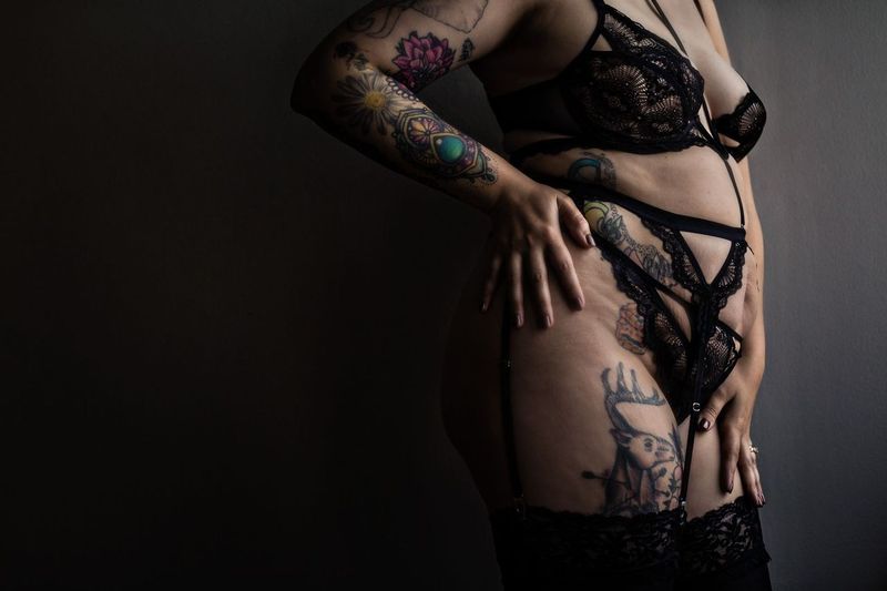 A woman wearing black lace lingerie is posing against a dark background. Their body is adorned with various colorful tattoos on the arms and abdomen.