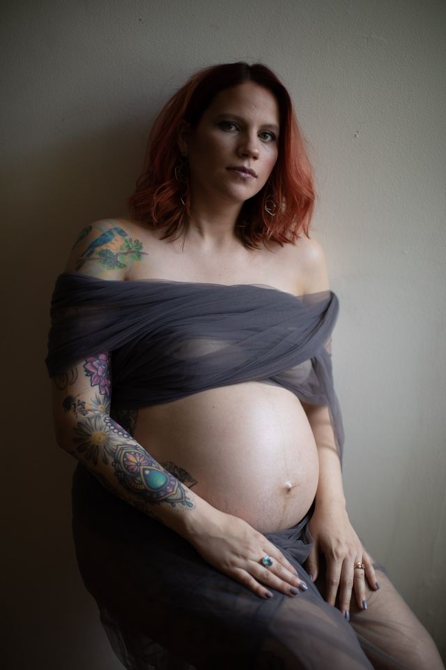A pregnant woman with red hair sits against a light wall. Shes wrapped in a sheer gray fabric, showcasing her tattooed arm. Her expression is calm and contemplative.