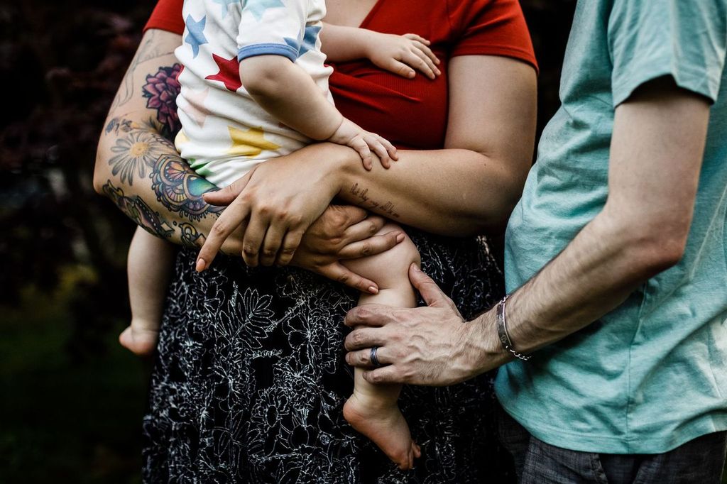 A person with a colorful arm tattoo holds a baby wearing a star-patterned outfit. Another person, wearing a mint green shirt with a ring on their hand, stands close, touching the babys leg gently. The background is dark and blurred.