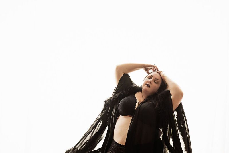 A woman poses confidently in black lingerie and a flowing sheer robe against a bright white background. They have their hands on their head with a relaxed expression, emphasizing empowerment and body positivity.