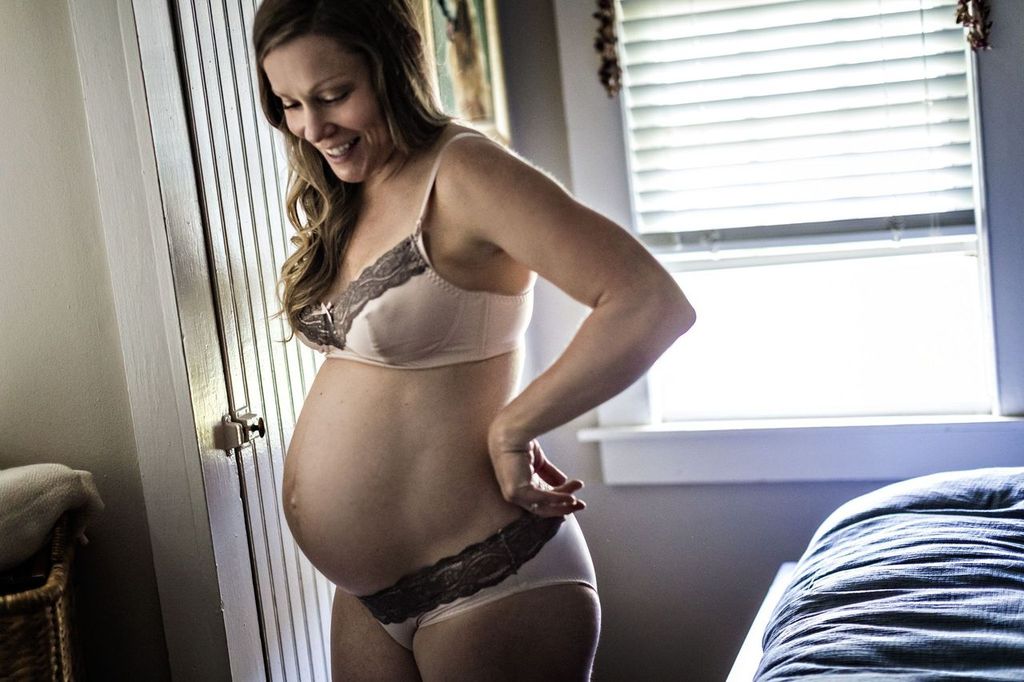 A pregnant woman smiling, standing in a room beside a bed. She is wearing lingerie and gently touching her belly. Sunlight filters through a window with blinds, creating a warm atmosphere.