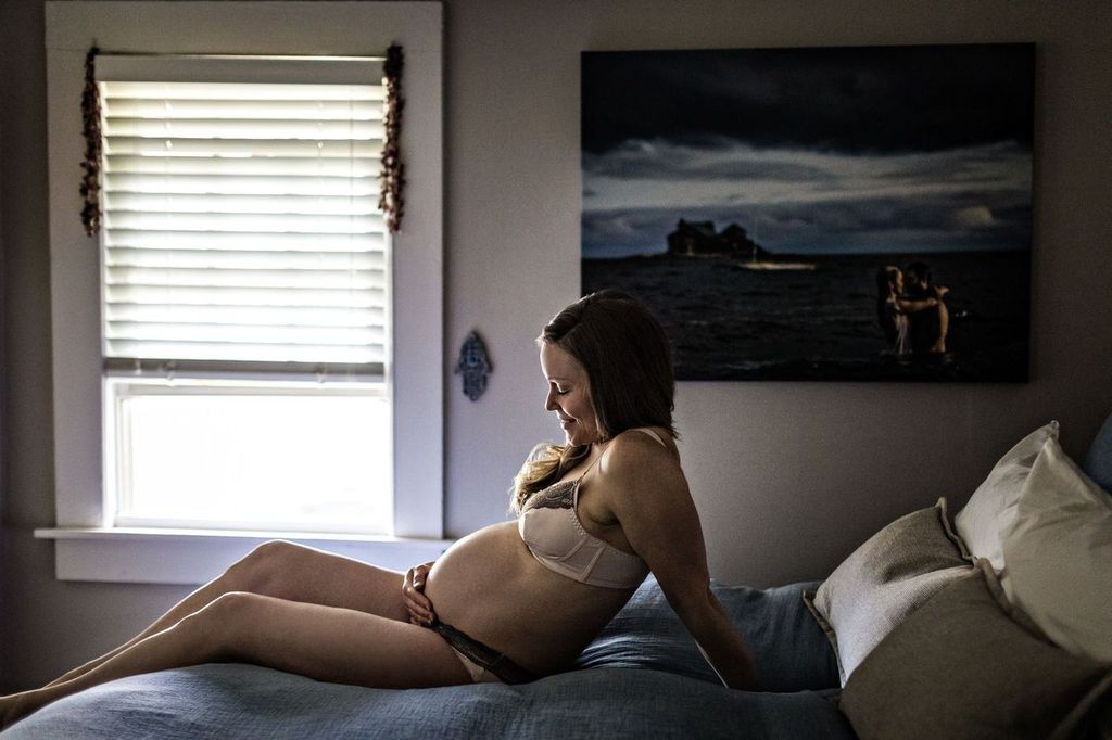 A pregnant woman wearing a bra and shorts sits on a bed in a softly lit room, gently touching her belly. A window with blinds and a framed seaside photograph on the wall are in the background.