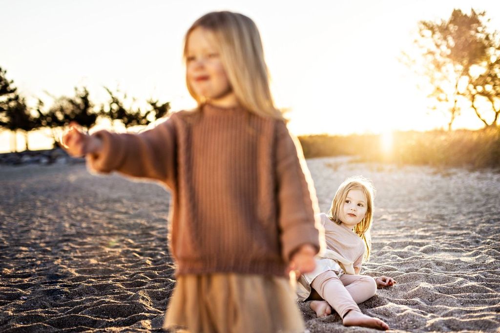 Two young children with long hair on a sandy beach. One stands in the foreground wearing a brown sweater, reaching out, while the other sits on the sand in a light outfit, gazing at the first child. Sunlight glows in the background.