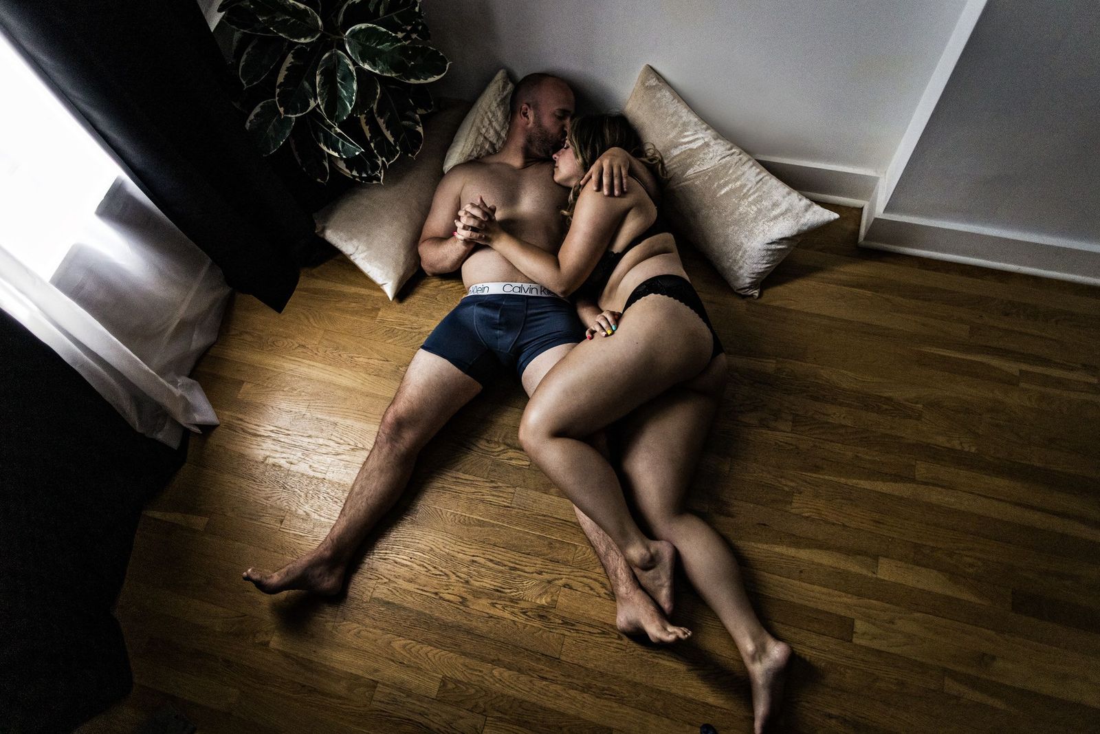 A couple lies on a wooden floor, holding hands and cuddling. They are surrounded by soft cushions, and a plant is visible nearby. Light streams in from a window, creating a peaceful and intimate atmosphere.