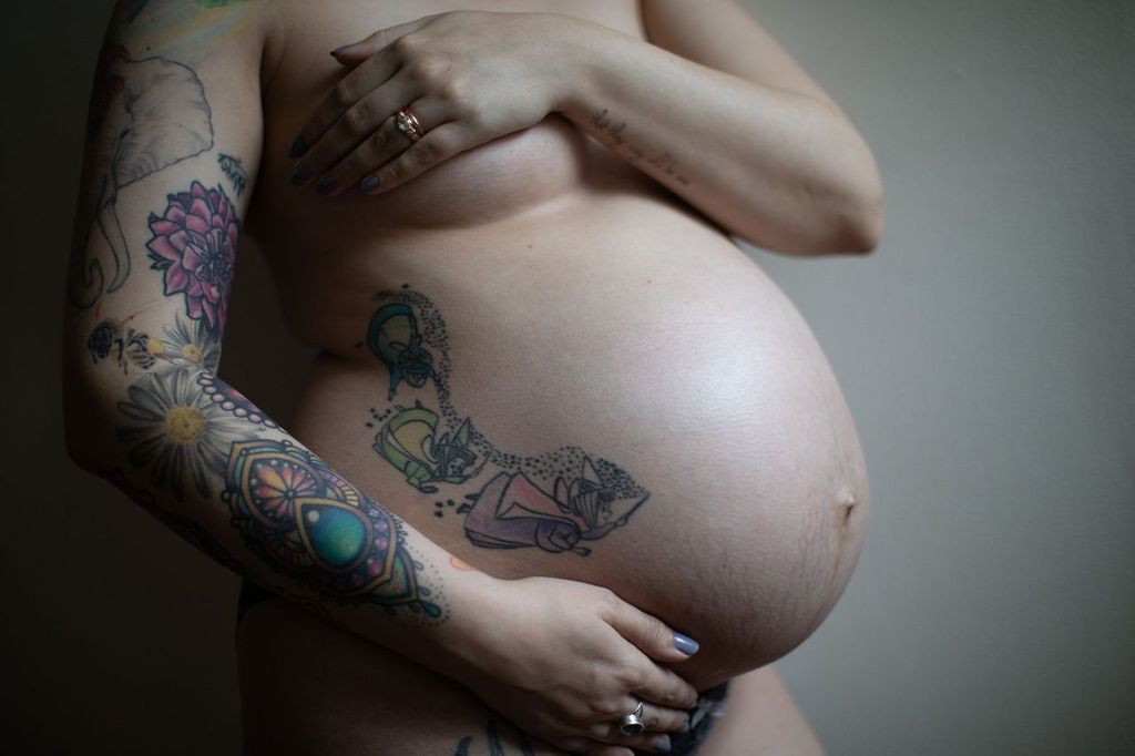 A pregnant woman stands against a plain background, showing a large, tattooed pregnant belly. The arm and side are also covered with colorful tattoos, including floral and abstract designs. Hands rest gently on the belly.