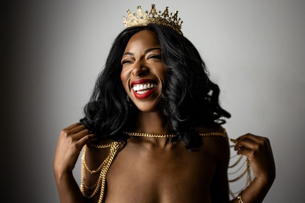A woman with long black hair and bright red lipstick is smiling broadly. She wears a gold crown and holds golden chains draped over her shoulders against a plain background.