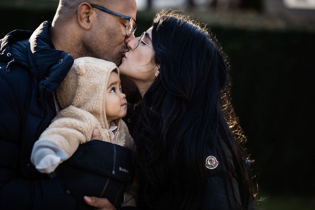 A couple wearing glasses kisses while holding a baby. The man holds the baby, who is dressed in a fluffy hooded outfit, as the woman leans in. They are outdoors, with a blurred green background.