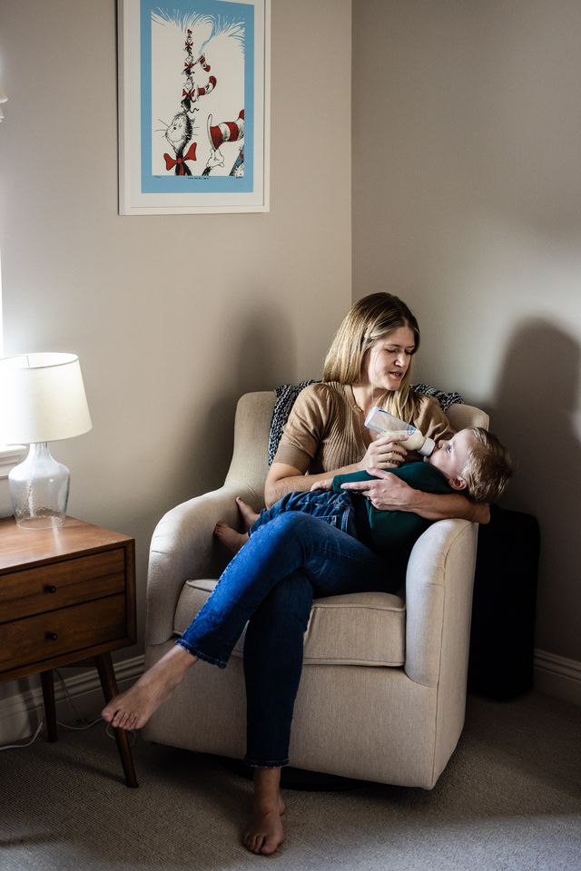 A woman sits in a beige armchair feeding a baby with a blue bottle. She is wearing a brown top and jeans. A lamp and wooden side table are next to her. A framed illustration of the Cat in the Hat hangs on the wall behind them.