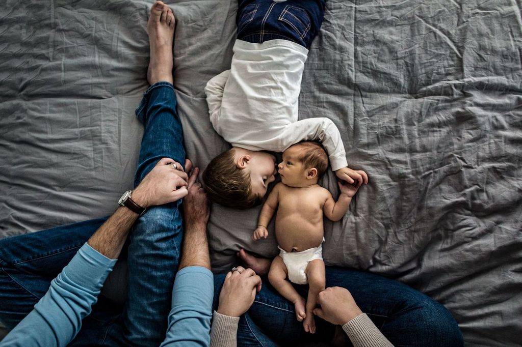Two adults sit on a bed facing two children. One child, wearing a white shirt, cuddles next to a baby in a diaper. Their legs and arms create a frame around the children on a gray bedspread.