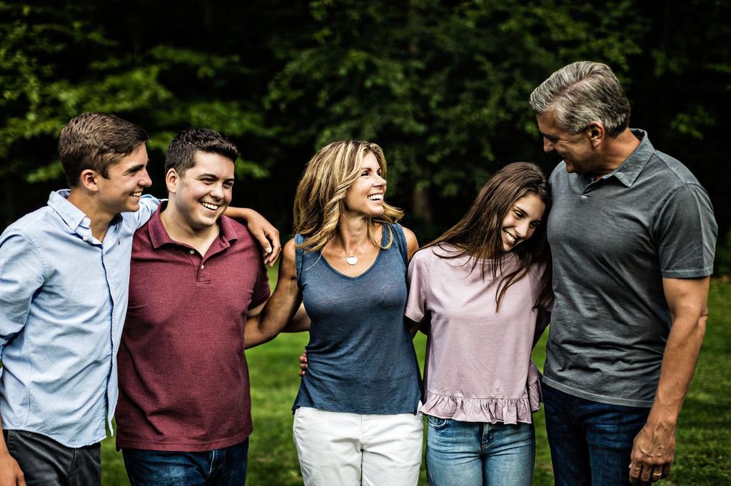 A family of five people stands together outdoors, smiling and with their arms around each other. They are casually dressed and appear to be enjoying each others company. Trees and grass are in the background.