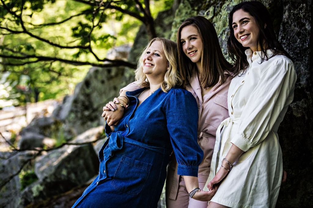 Three women stand together in an outdoor setting, smiling and leaning against a rock surface. They are wearing casual dresses in blue, light pink, and white. Trees and rocks are visible in the background, creating a natural ambiance.