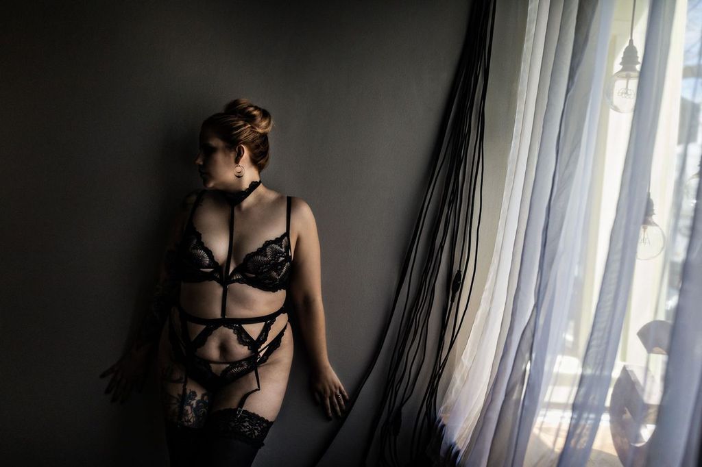 A woman in black lace lingerie stands by a window with sheer curtains. She leans against a wall, gazing toward the light. Her hair is styled in a bun, and tattoos are visible on her arm and thigh. The room is dimly lit.