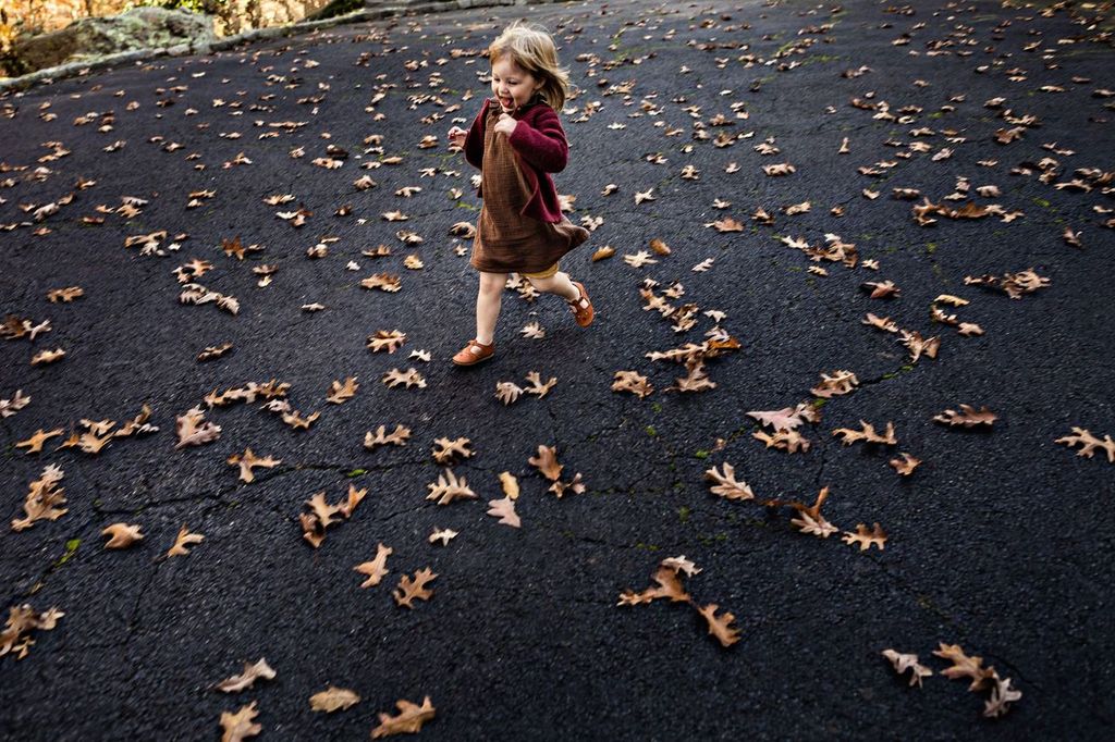 A young child in a brown dress and maroon cardigan joyfully runs through a scattering of brown leaves on a paved surface. The child is smiling and appears to be playing in the midst of autumn foliage.