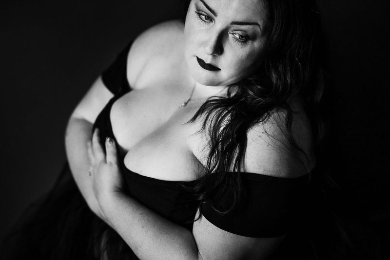 Black and white portrait of a woman with long hair, wearing an off-shoulder top. She gazes thoughtfully to the side, her right arm crossed over her chest, creating a contemplative and introspective atmosphere.